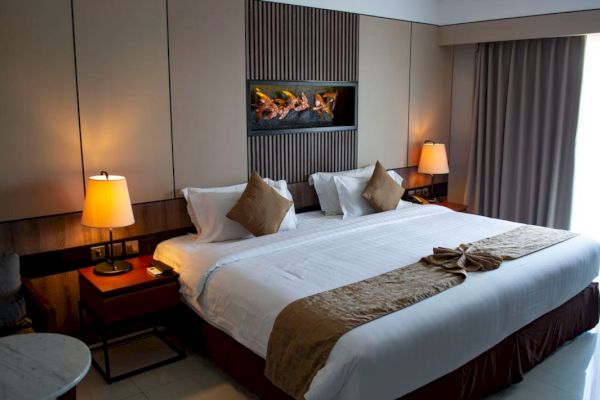 The image depicts a modern hotel room with a large, neatly made bed, two side tables with lamps, and a window with heavy curtains.