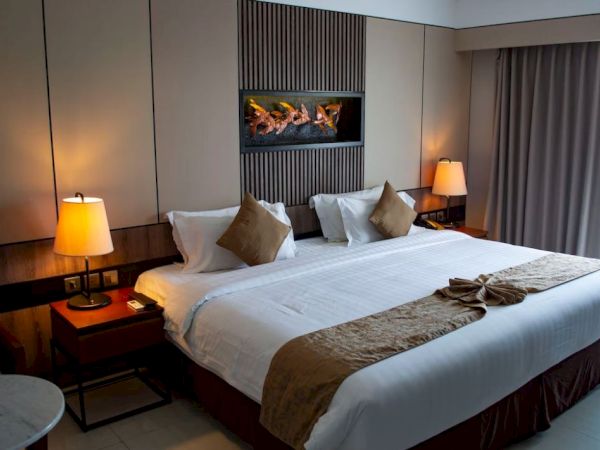 The image depicts a modern hotel room with a large, neatly made bed, two side tables with lamps, and a window with heavy curtains.