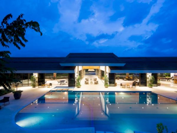 The image shows a luxurious outdoor swimming pool area at dusk, with modern architecture, deck chairs, and a well-lit building in the background.