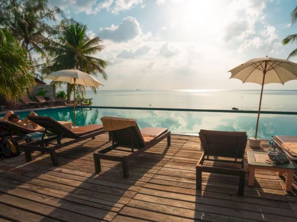 The image shows a tranquil poolside setting with lounge chairs, umbrellas, palm trees, and a view of the ocean under a partly cloudy sky.