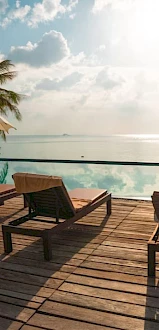 A serene poolside scene with lounge chairs, umbrellas, and a view of the ocean at sunset or sunrise, surrounded by tropical palm trees.