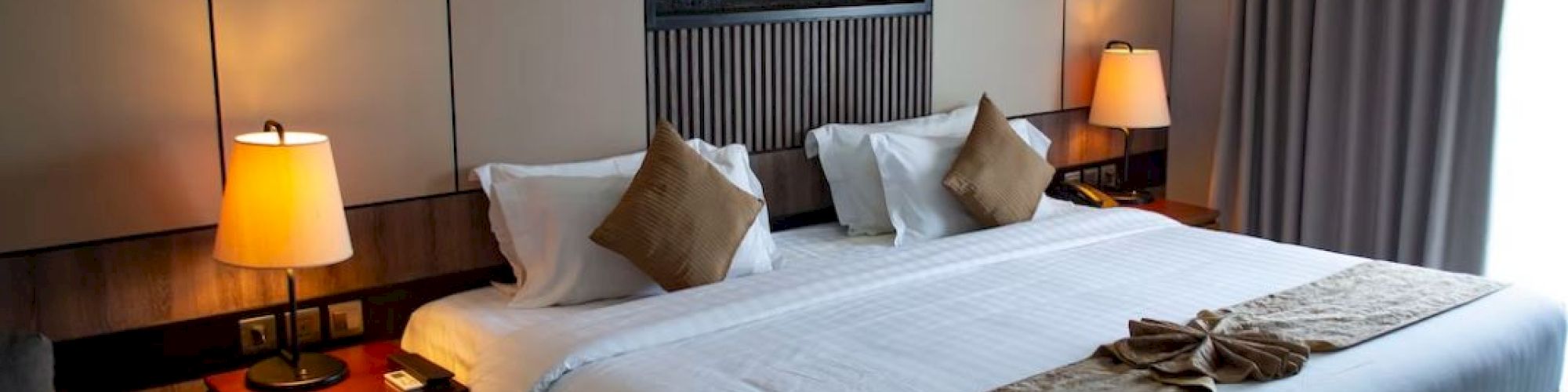 A neatly made bed with pillows and blanket accents, bedside tables with lamps, a small round table, and a decorative wall piece is shown.