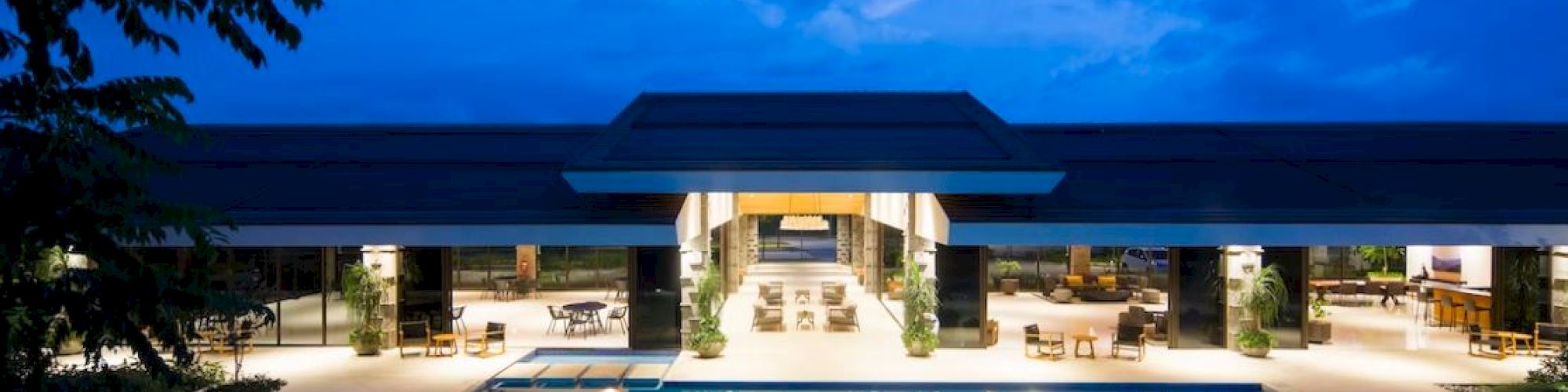 A modern building with an illuminated pool at night, featuring open lounge areas and outdoor seating, set against a vibrant evening sky.