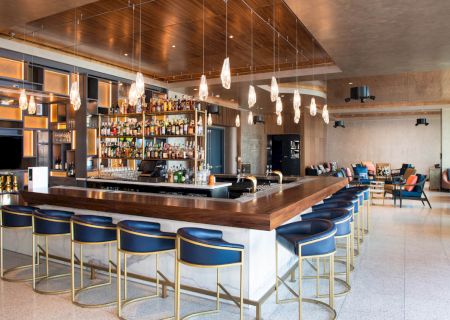 The image shows a modern bar with a wooden counter, blue cushioned stools, hanging lights, a variety of alcohol bottles, and a lounge area in the background.