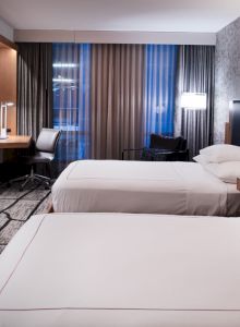 The image shows a modern hotel room with two double beds, a wall-mounted TV, a desk with a chair, and a window with curtains.