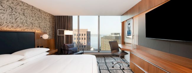 A modern hotel room with a large bed, wide TV, wooden furniture, desk, and a window with a city view and some buildings outside, as well as an armchair.