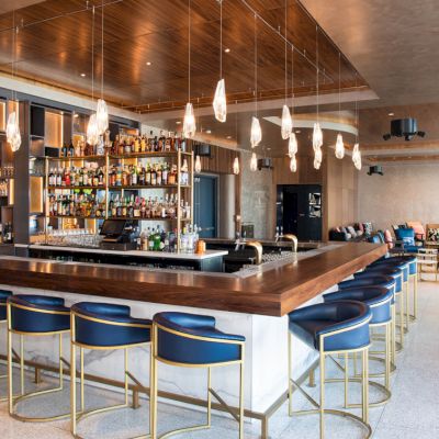 This image shows a modern bar with stylish lighting, a well-stocked array of bottles, and blue cushioned barstools, within a sleek, contemporary setting.