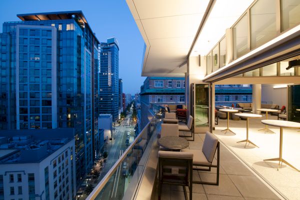 A modern balcony overlooks a city street with tall buildings. The balcony is furnished with tables and chairs, and the view is during dusk.