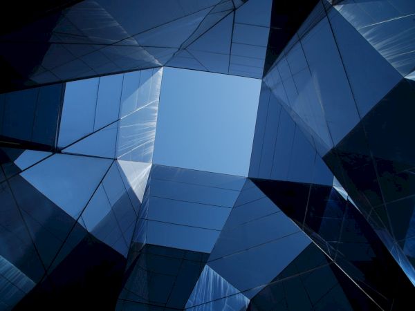 The image shows an upward view of a modern glass building with reflective surfaces forming geometric patterns, framing a blue sky.