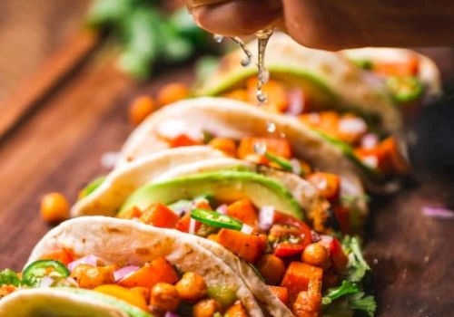 The image shows a hand squeezing lime juice over a row of tacos filled with ingredients like chickpeas, diced vegetables, cilantro, and avocado slices.