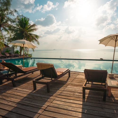 A serene beach scene with lounge chairs, umbrellas, palm trees, and an infinity pool overlooking the ocean under a partly cloudy sky.