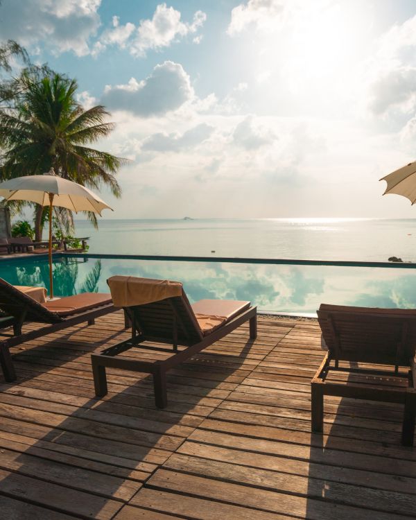 A serene beach scene with lounge chairs, umbrellas, palm trees, and an infinity pool overlooking the ocean under a partly cloudy sky.