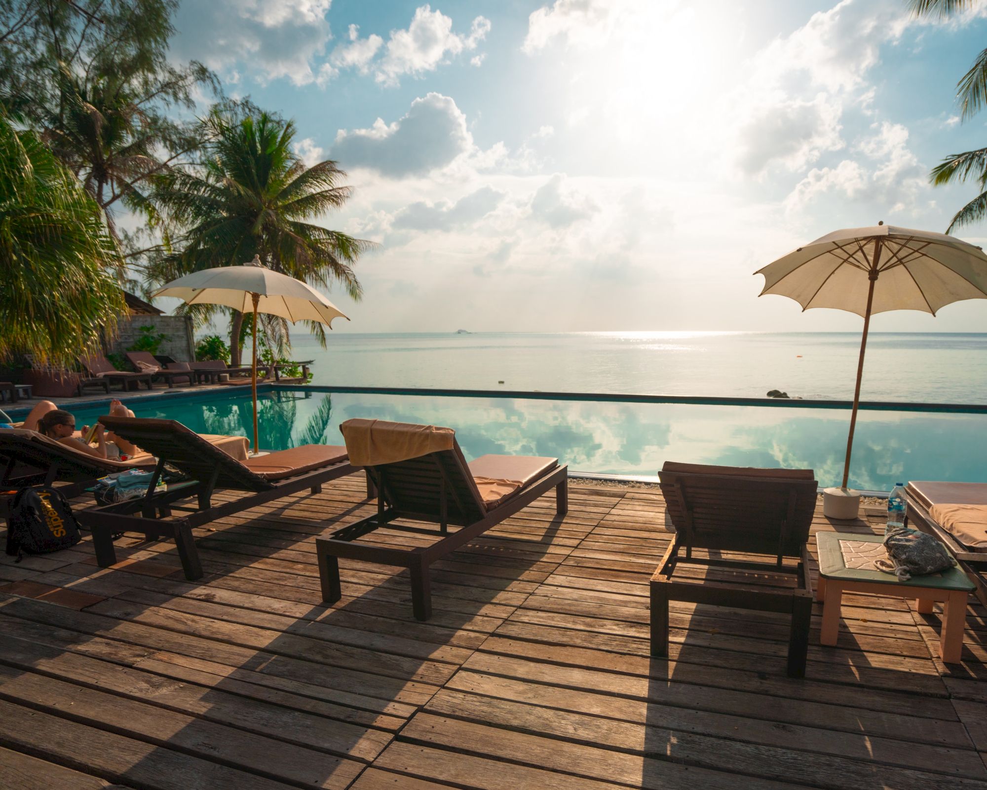 A serene tropical scene with lounge chairs and umbrellas by a wooden deck, overlooking a calm infinity pool and ocean with a bright sun in the sky.