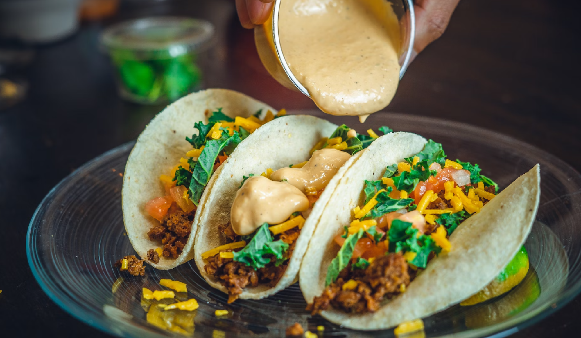 A hand is pouring sauce onto three tacos filled with ground meat, cheese, and vegetables on a glass plate. There's a small container in the background.