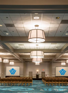 The image shows a conference room with rows of chairs facing two projection screens and a podium, under several large hanging lights.