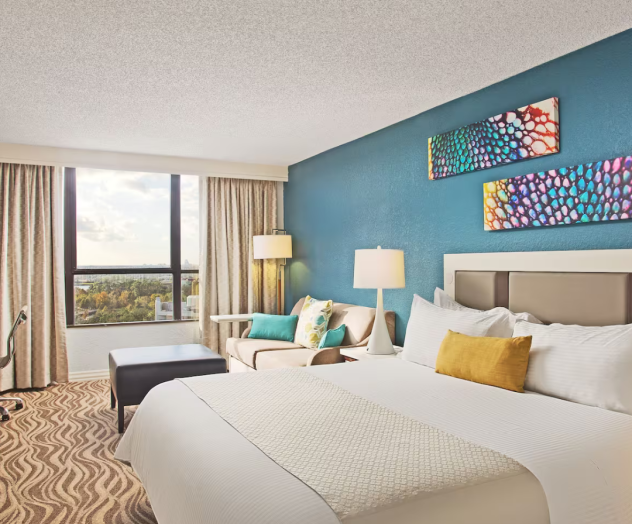 A modern hotel room with a bed, colorful artwork, a chair, a side table lamp, and a large window with a view.