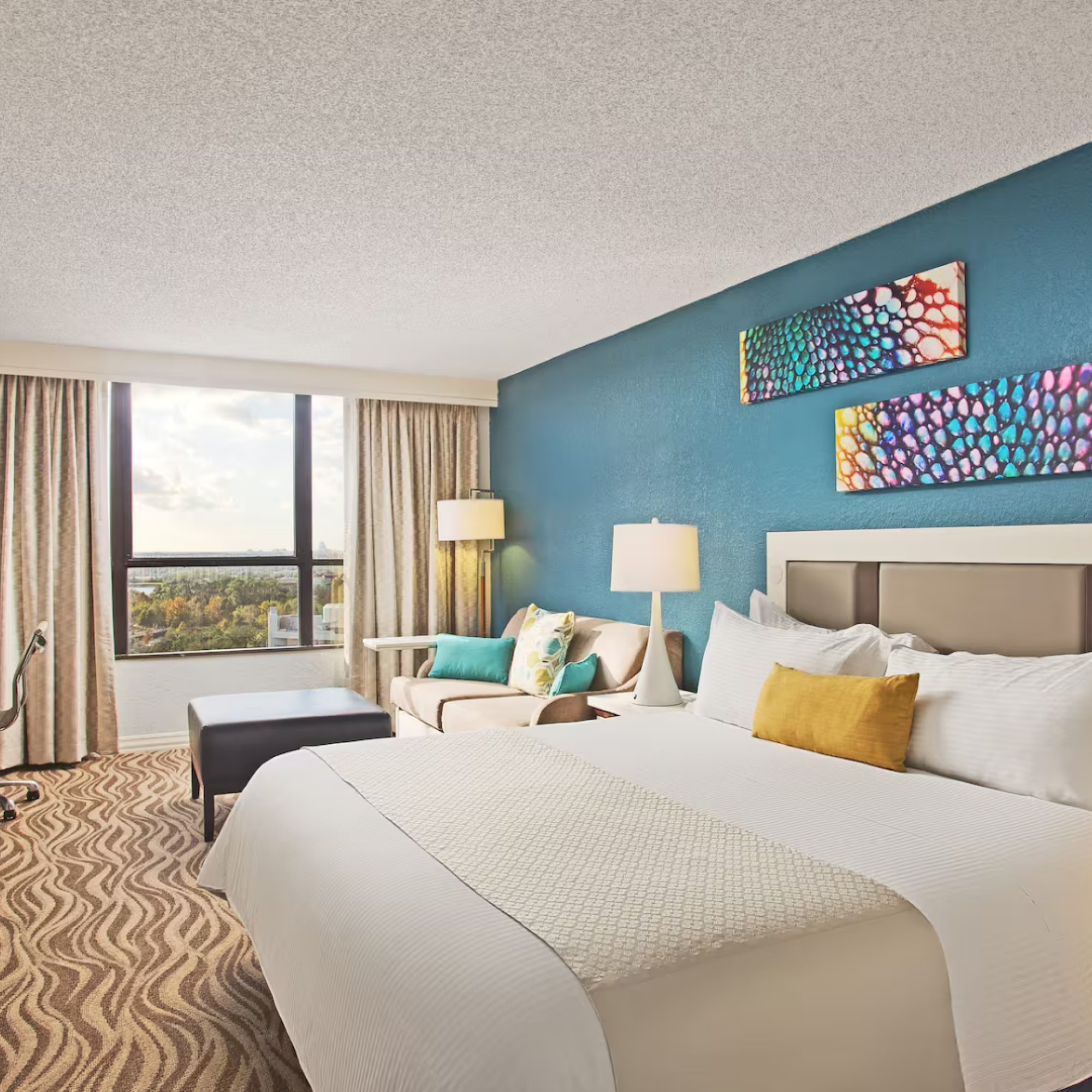 A modern hotel room with a large bed, colorful artwork, armchair, desk, and window view with curtains. It has a blue accent wall and floor lamp.