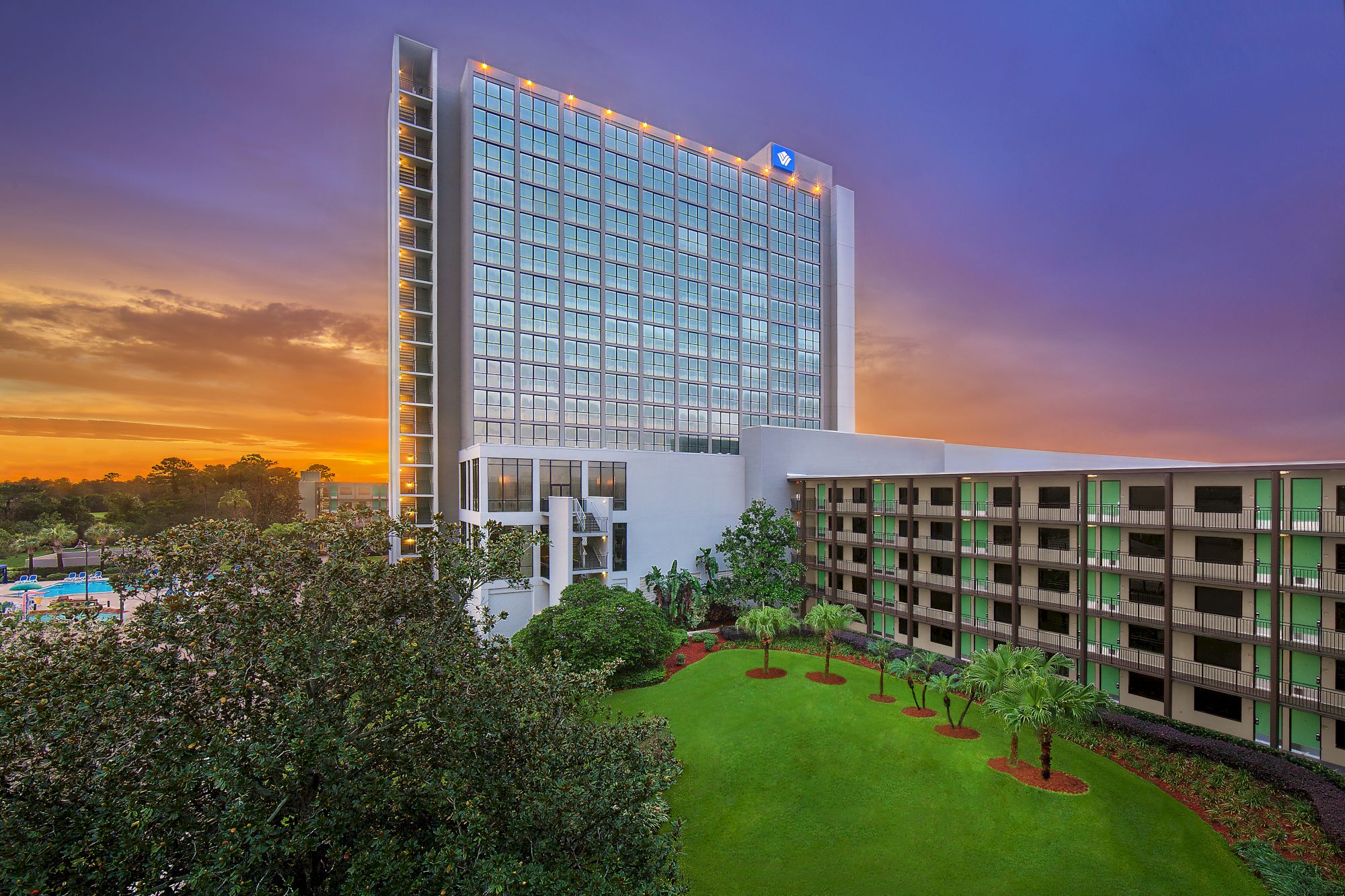 A tall hotel building with a smaller adjacent structure, surrounded by greenery and a beautiful sunset sky is shown in the image.