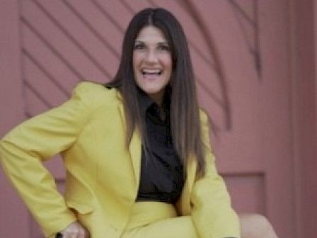 A woman dressed in a yellow suit and black blouse is posing outdoors, smiling at the camera, with a pink door in the background.