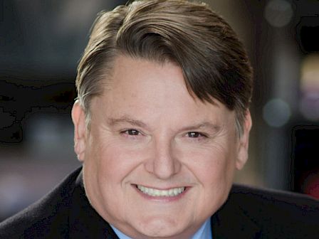 A person wearing a suit and a tie with a pattern of smiling faces. The background is blurred, focusing on the individual's face.