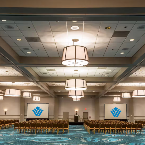 The image shows a conference room with rows of chairs, two projection screens displaying logos, and large ceiling lights hanging from the ceiling.