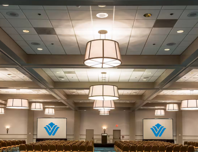 The image shows a conference room with rows of chairs, two projection screens displaying logos, and large ceiling lights hanging from the ceiling.