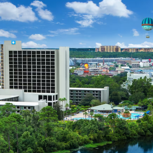 A tall hotel stands surrounded by lush greenery and a river, with a cityscape and hot air balloon visible in the background under a blue sky.