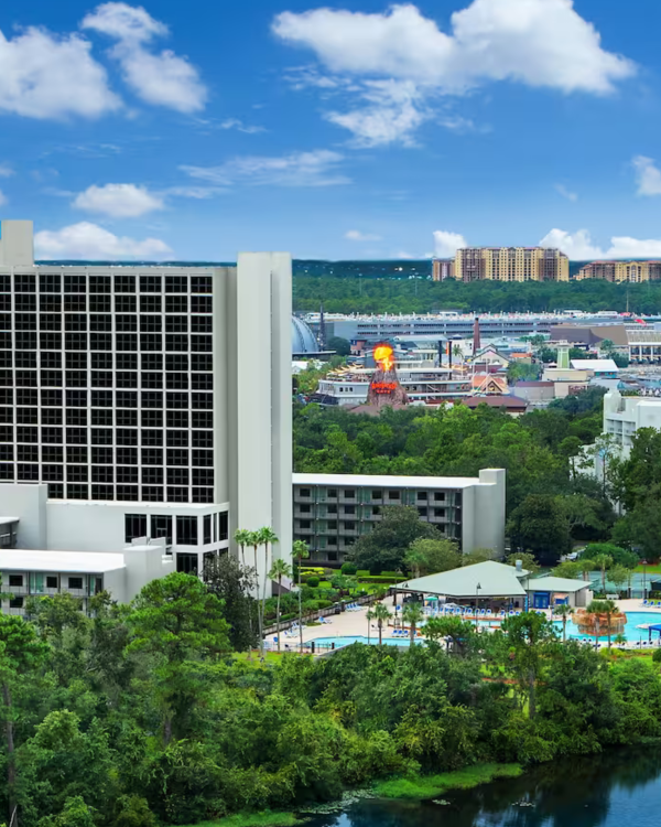 A tall hotel stands surrounded by lush greenery and a river, with a cityscape and hot air balloon visible in the background under a blue sky.