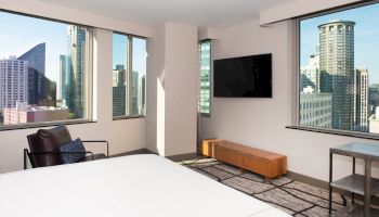 This image shows a modern hotel room with a bed, armchair, wall-mounted TV, and large windows with a cityscape view, ending the sentence.