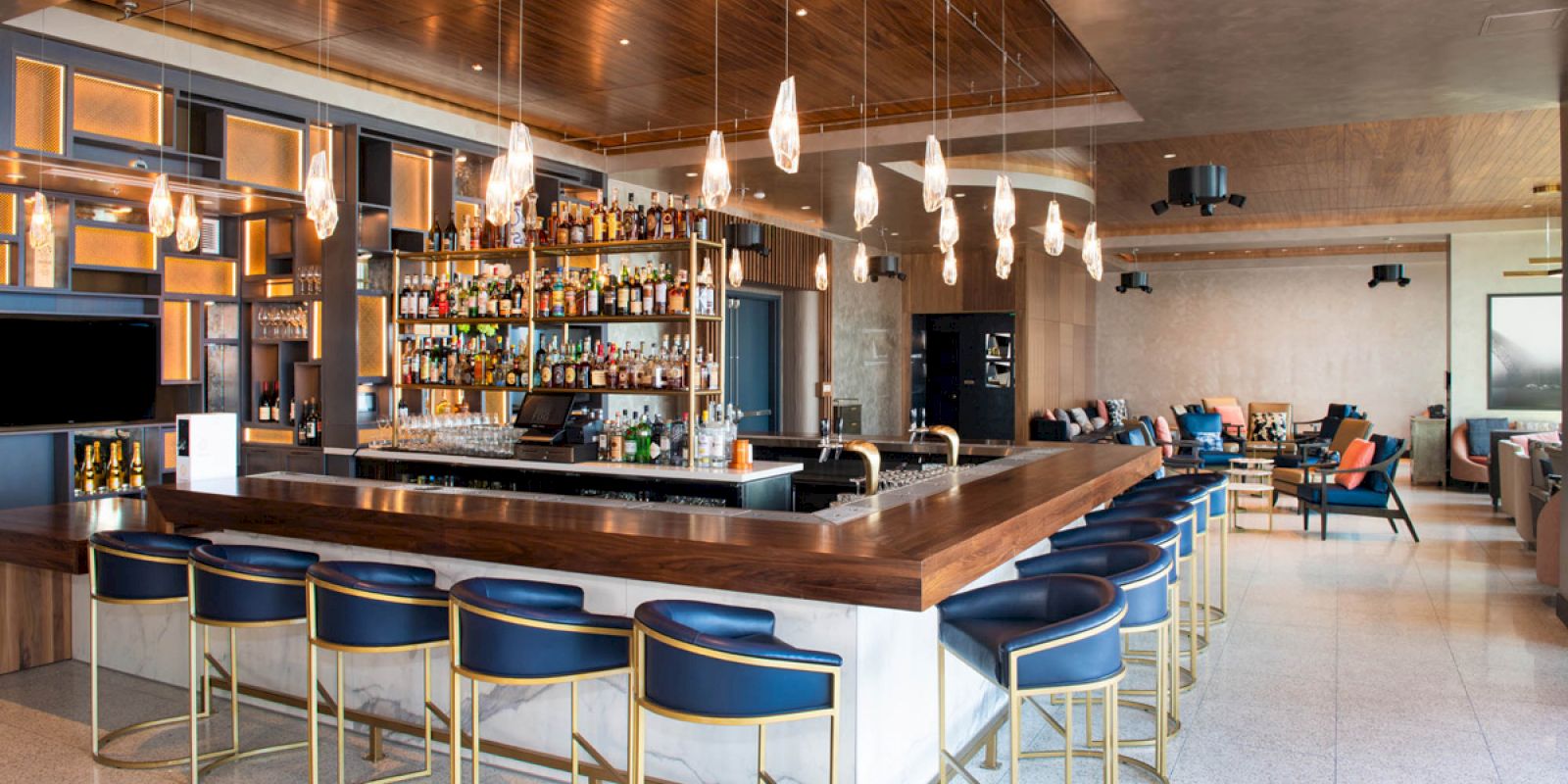 A modern bar with a wooden counter, blue cushioned barstools, multiple hanging lights, and a well-stocked liquor display can be seen.