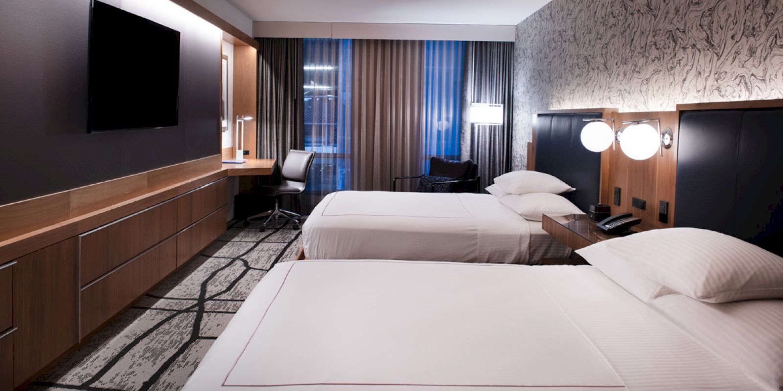This image shows a modern hotel room with two neatly made twin beds, a flat-screen TV, a desk with a chair, and decorative lighting.