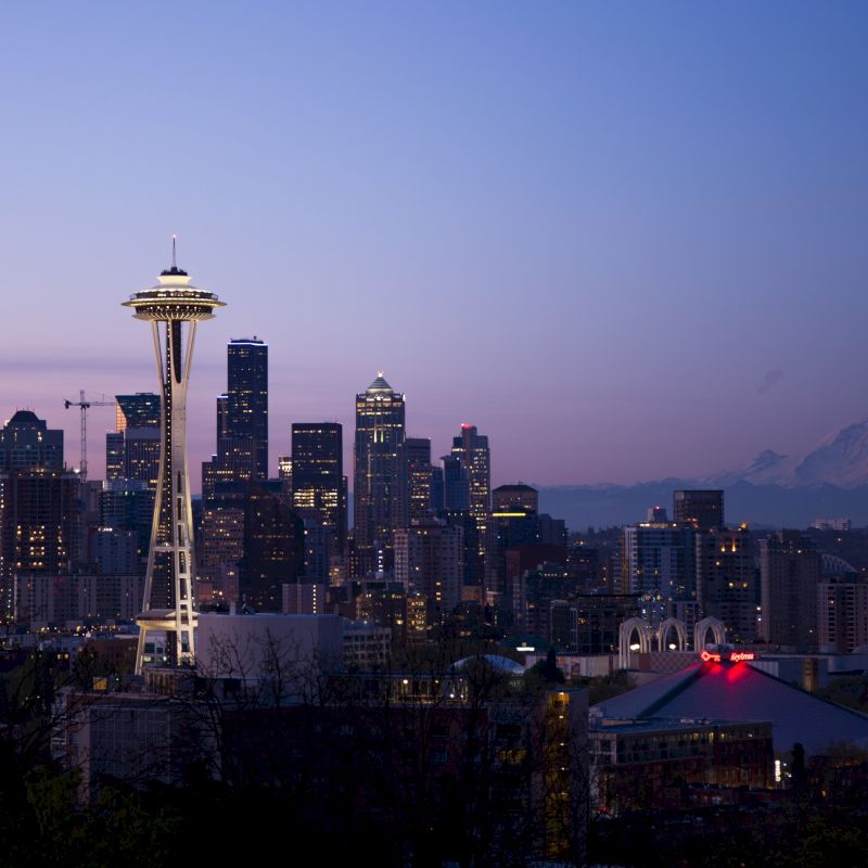 The image shows a twilight view of Seattle's skyline, highlighted by the Space Needle, with Mount Rainier visible in the background.