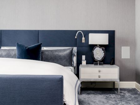 A modern bedroom features a blue upholstered headboard, white bedside table with a lamp and decor, grey walls, and a neatly made bed.