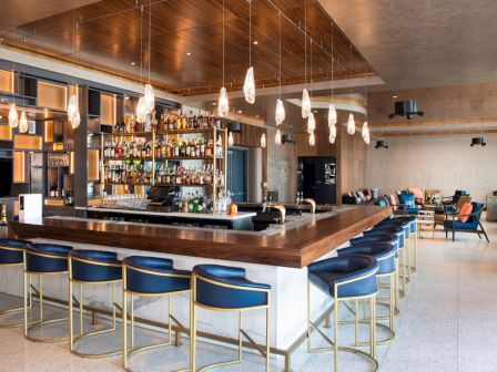 The image depicts a modern bar with a large counter, blue cushioned high stools, overhead pendant lights, and a well-stocked bar area.