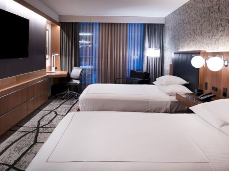 The image shows a modern hotel room with two beds, a desk, a TV, and stylish decor.