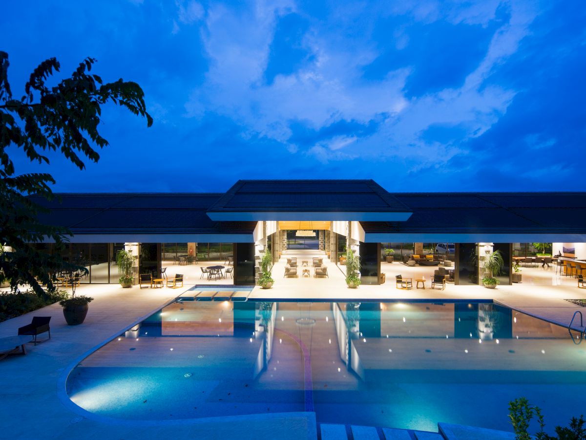 A luxurious building with open floor plan and illuminated pool at dusk, surrounded by trees and outdoor seating areas, under a blue sky.