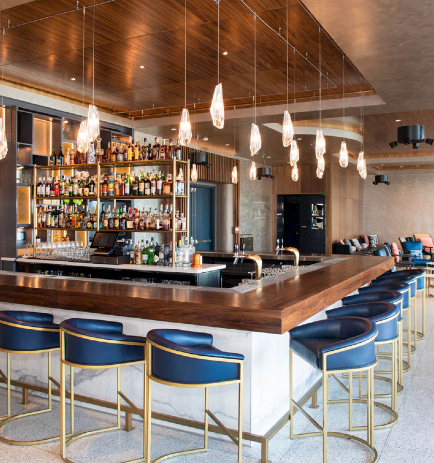 A modern bar with a wooden counter, blue and gold bar stools, various bottles on shelves, and ambient lighting in a stylish interior setting.