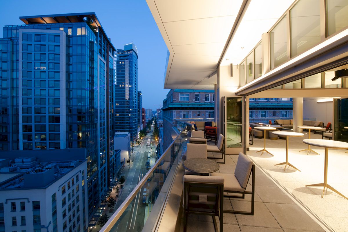 This image shows a modern balcony with outdoor furniture, overlooking a cityscape with tall buildings in the evening sky.