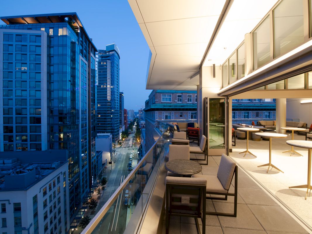 This image shows a modern balcony with outdoor furniture, overlooking a cityscape with tall buildings in the evening sky.
