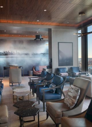 A modern lounge room featuring comfortable chairs, tables, and large windows with a scenic view of a waterfront and skyline, ending the sentence.