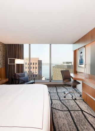 A modern hotel room features a large bed, a desk with a chair, a flat-screen TV, and a window with a city view.