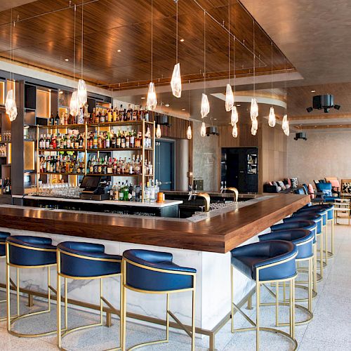 The image shows a modern bar with blue and gold barstools, a wooden countertop, and various bottles displayed behind the bar.