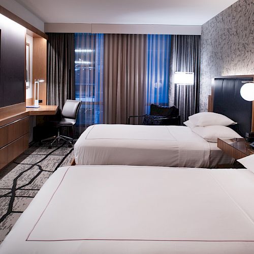 The image shows a modern hotel room with two beds, a wall-mounted TV, desk, chair, lamps, and a window with curtains.