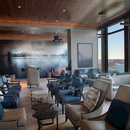 The image shows a modern lounge with comfortable seating, large windows offering a waterfront view, and a wall artwork featuring city skyline.