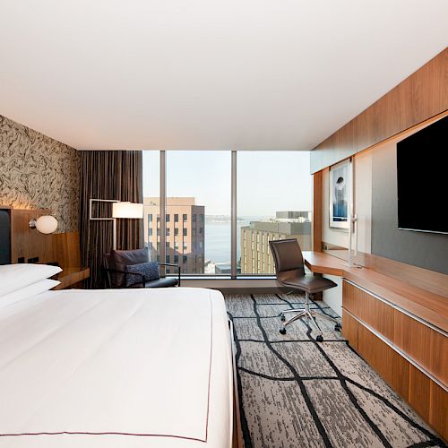A modern hotel room with a large bed, TV, desk, and chair. The room has a city view from floor-to-ceiling windows and contemporary decor, ending the sentence.