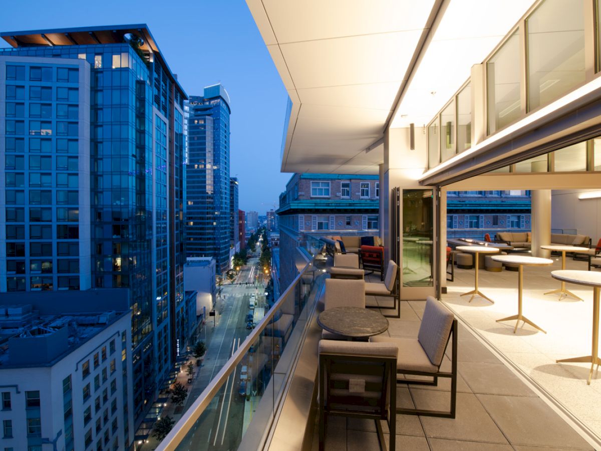 A modern city view from an elevated outdoor balcony at dusk, showing high-rise buildings and a furnished terrace with seating and tables.