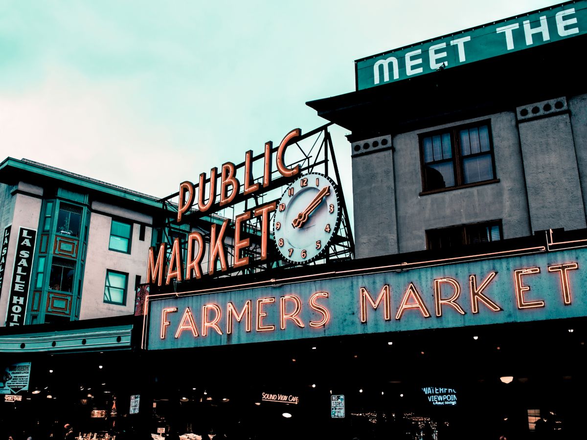 A neon sign reading "Public Market" and "Farmers Market" is displayed on a building. A clock is integrated into the sign.