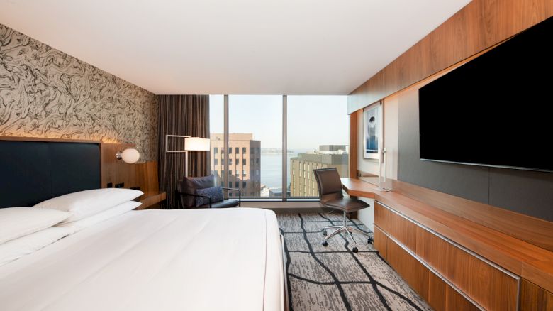 A modern hotel room featuring a bed, a large TV, a desk with a chair, and a window showing a cityscape.