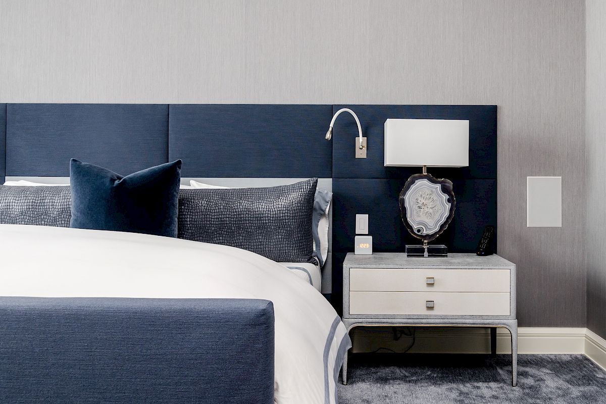 A modern bedroom features a blue upholstered headboard, matching pillows, white bedding, and a nightstand with a lamp and decorative item.