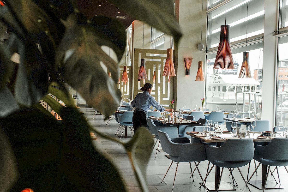A restaurant interior with modern decor, features pendant lights, neatly arranged tables, and a person preparing the dining area near large windows.
